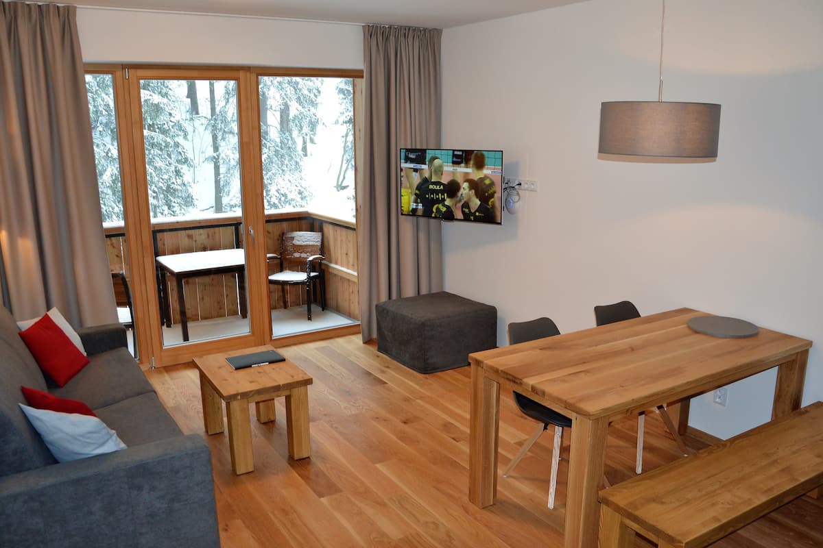 A modern living room with a wooden floor, grey sofa, wooden dining table, and a TV mounted on the wall.