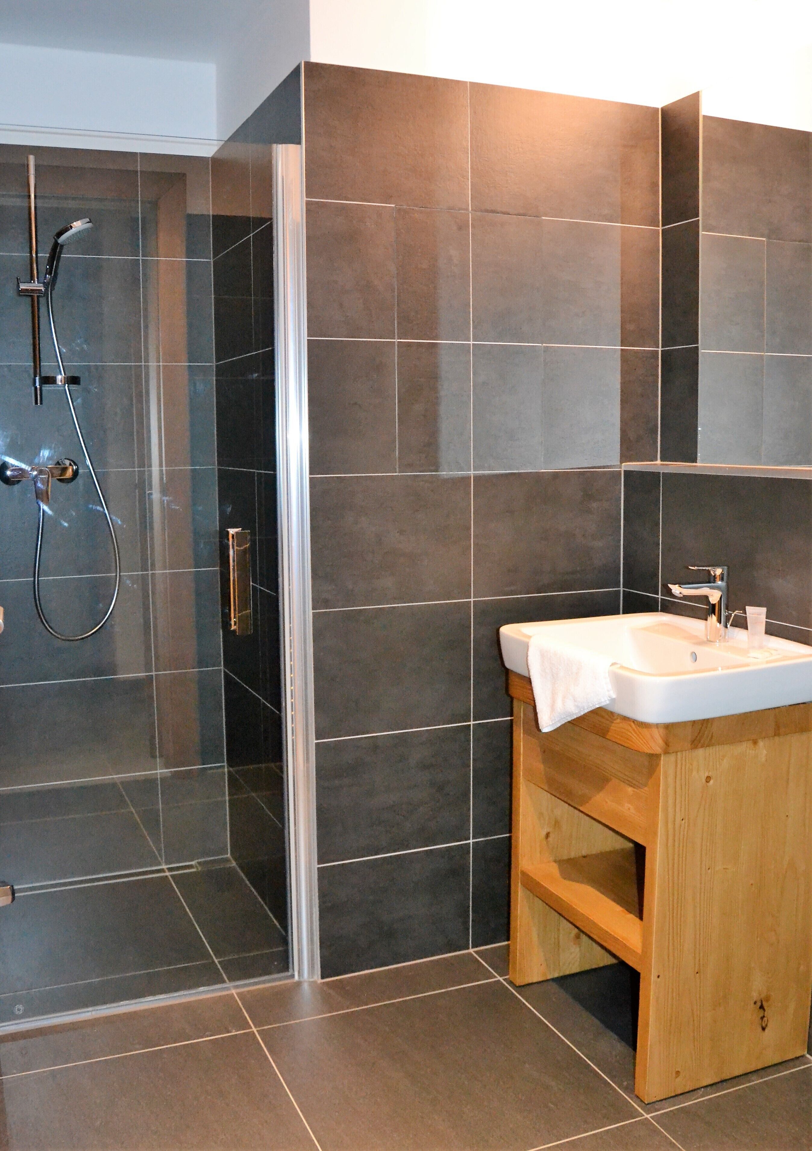A modern bathroom with a glass-enclosed shower, a wooden vanity, and dark tiled walls.