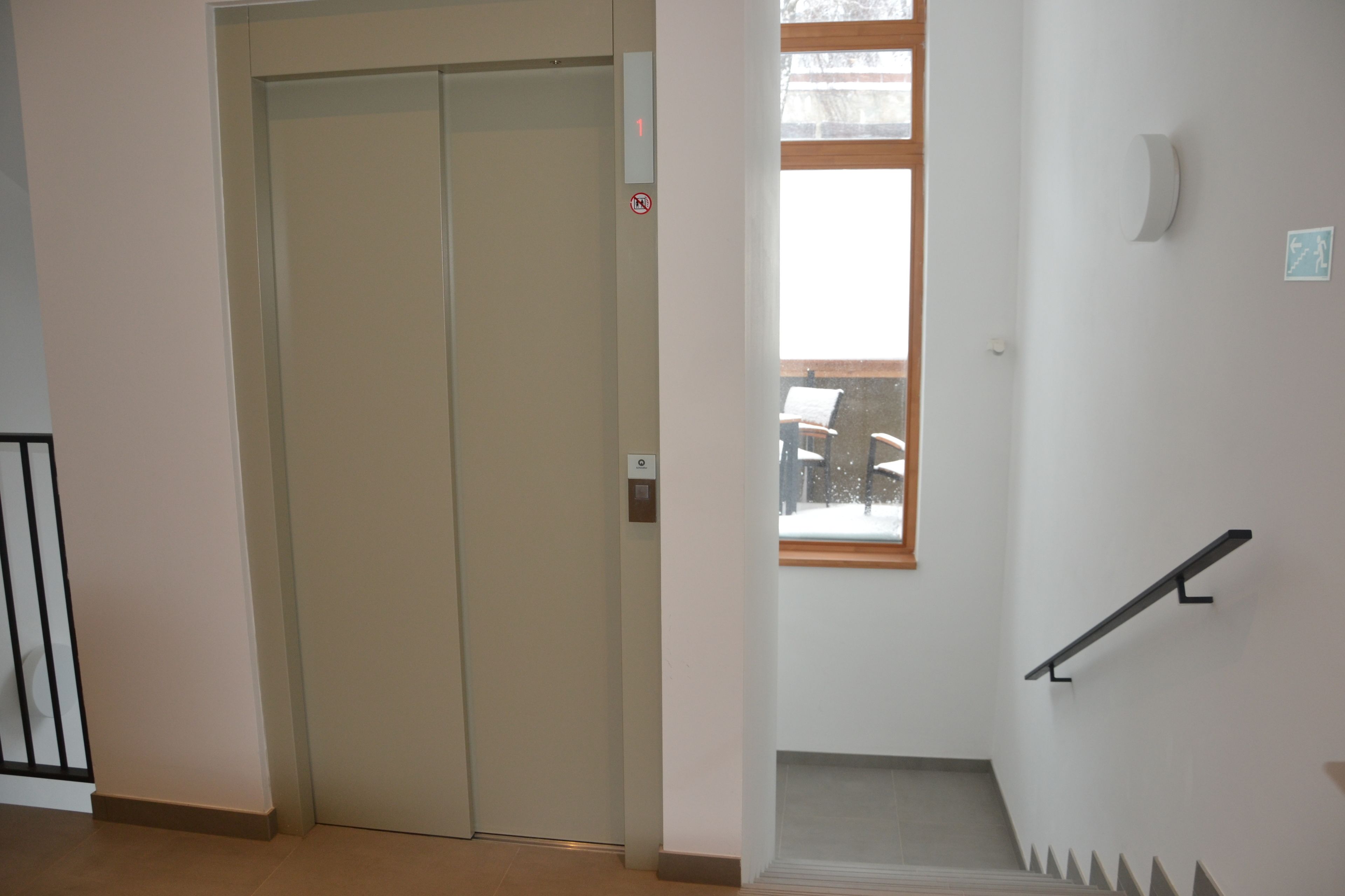 A staircase with a handrail, a door with a number sign, and a window with a view of outdoor furniture.