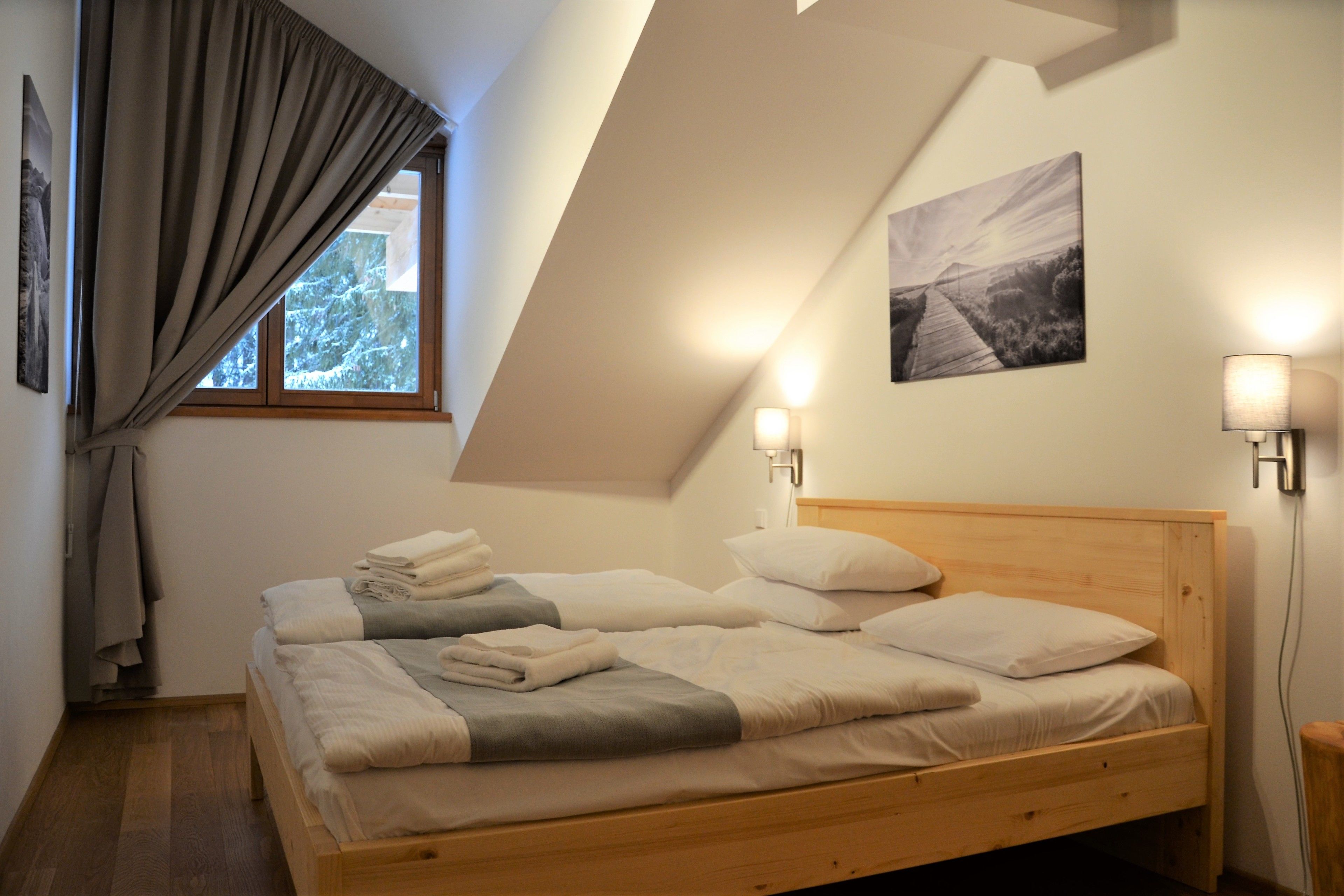 A bedroom with two beds, a wooden headboard, a window with curtains, wall-mounted lights, and a framed picture.