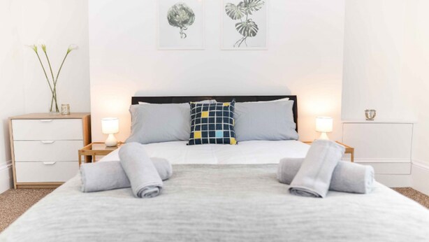 Una cama bien tendida con dos almohadas, una mesita de noche con una lámpara y una pequeña mesa con un florero.