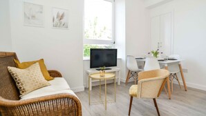 Una sala de estar con un sofá, televisor, mesa de comedor con sillas y ventana con vista a vegetación.