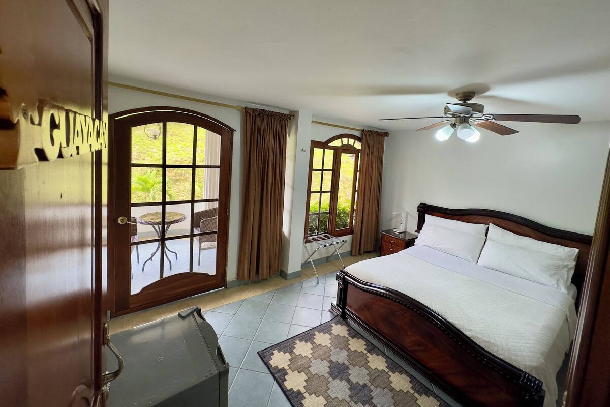 Ein Hotelzimmer mit einem Bett, einem Deckenventilator, einem Fenster mit Vorhängen, einem kleinen Tisch und Blick ins Freie.