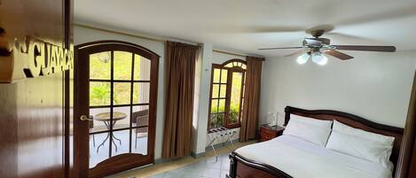 A hotel room with a bed, a ceiling fan, a window with curtains, a small table, and a view of the outdoors.