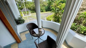 Un espace extérieur abrité, doté de deux fauteuils en osier et d’une petite table, entouré d’une balustrade blanche et offrant une vue sur un jardin.