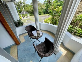 Un espacio al aire libre cubierto con dos sillones de mimbre y una mesita, rodeado por una barandilla blanca y vistas a un jardín.