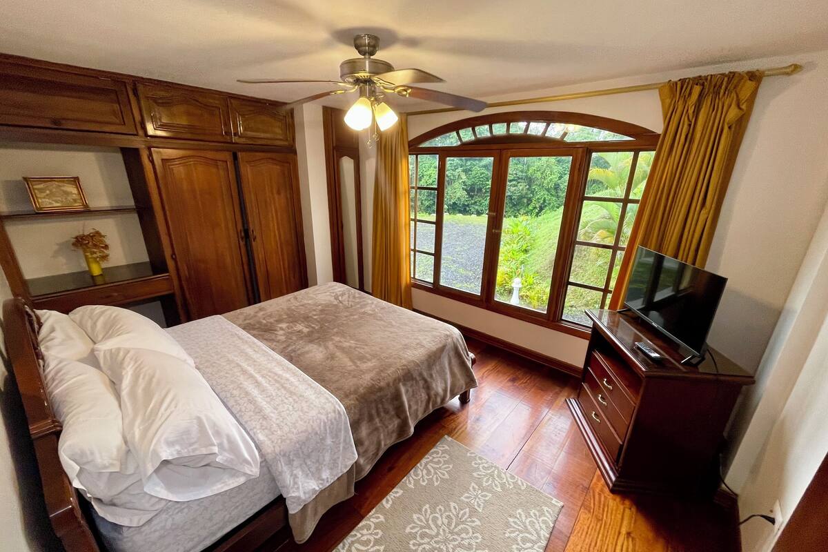 Ein Schlafzimmer mit Bett, Deckenventilator, Fernseher und einem Fenster mit Vorhängen.