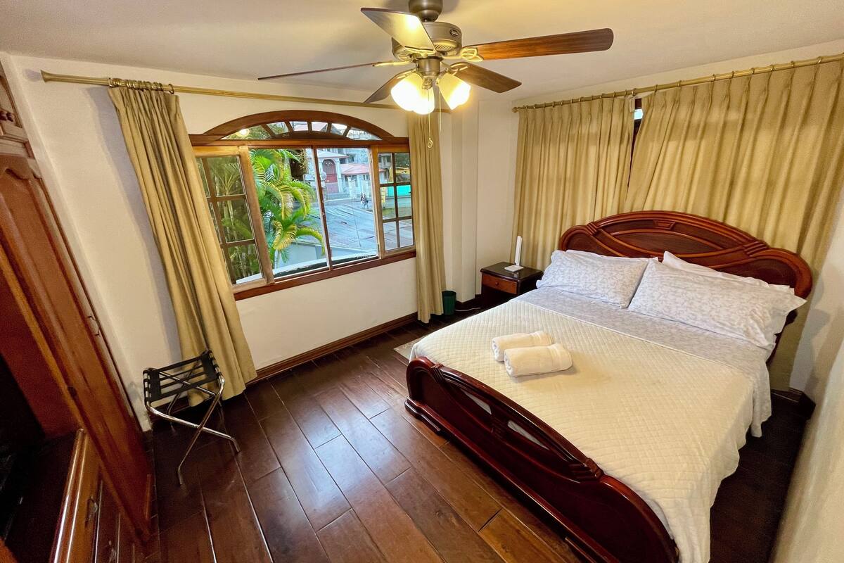 Ein Hotelzimmer mit einem hölzernen Bett, einem Deckenventilator, einem Fenster mit Vorhängen und Blick auf die Straße.