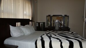 A hotel room with a large bed, a wooden headboard, a bedside table, a dresser, a mirror, and a window with curtains. - Premier Guest Lodge (Harare)