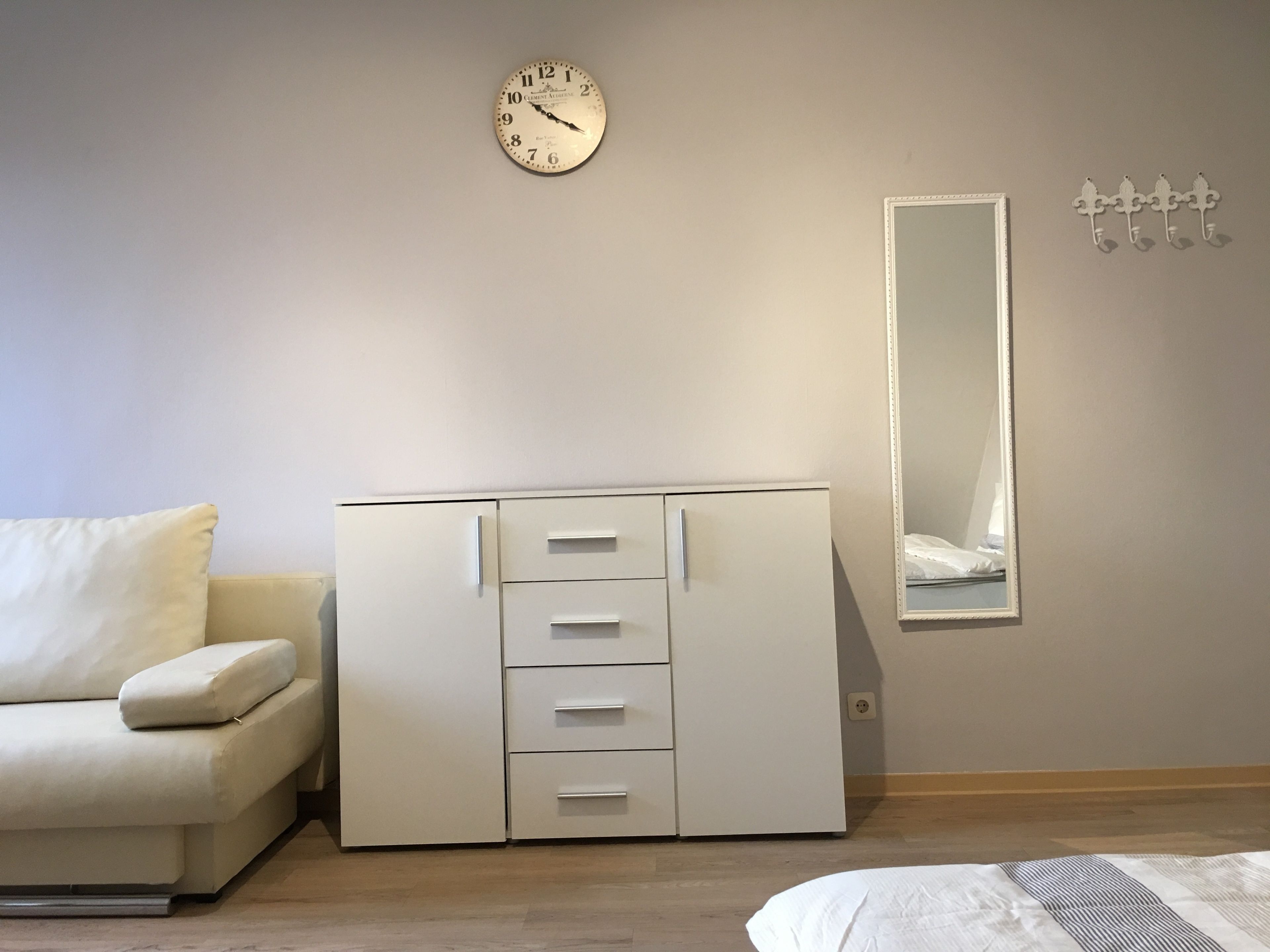 Una habitación minimalista con un sofá blanco, un armario blanco con cajones, un reloj de pared y un espejo.