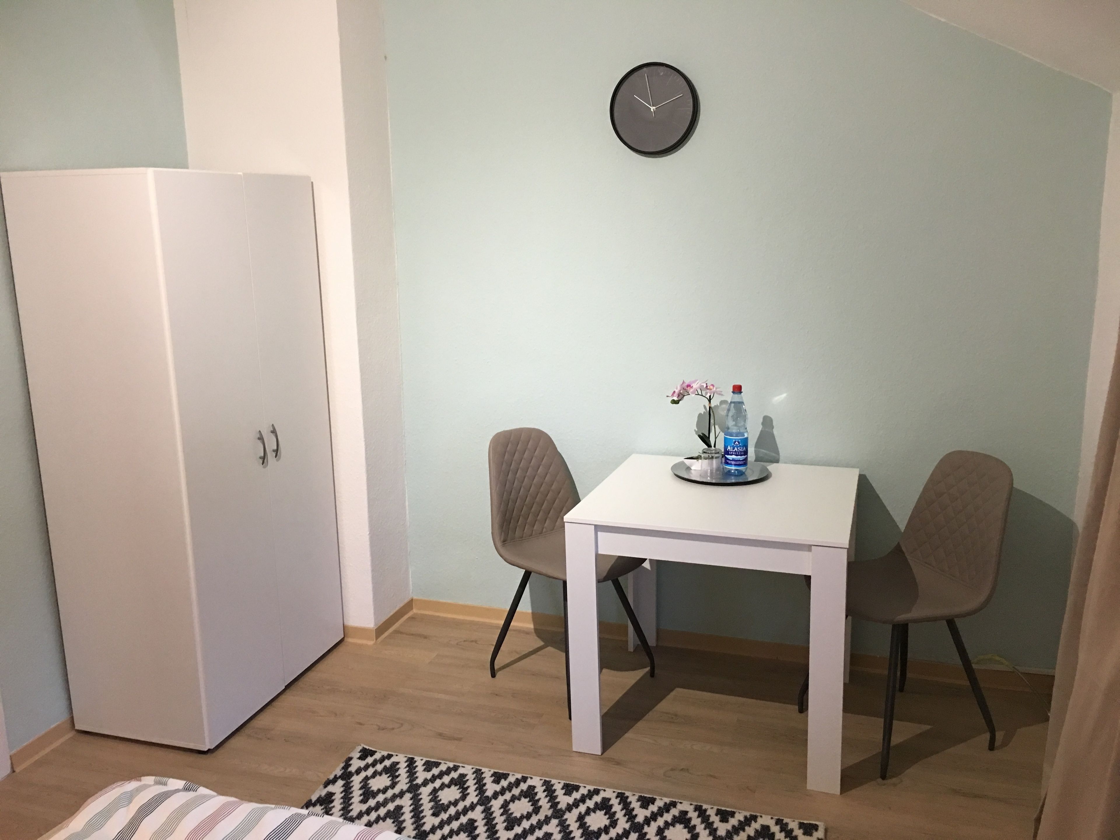 Una habitación pequeña y limpia con un armario blanco, una mesa redonda con dos sillas, un reloj en la pared y una botella de agua sobre la mesa.