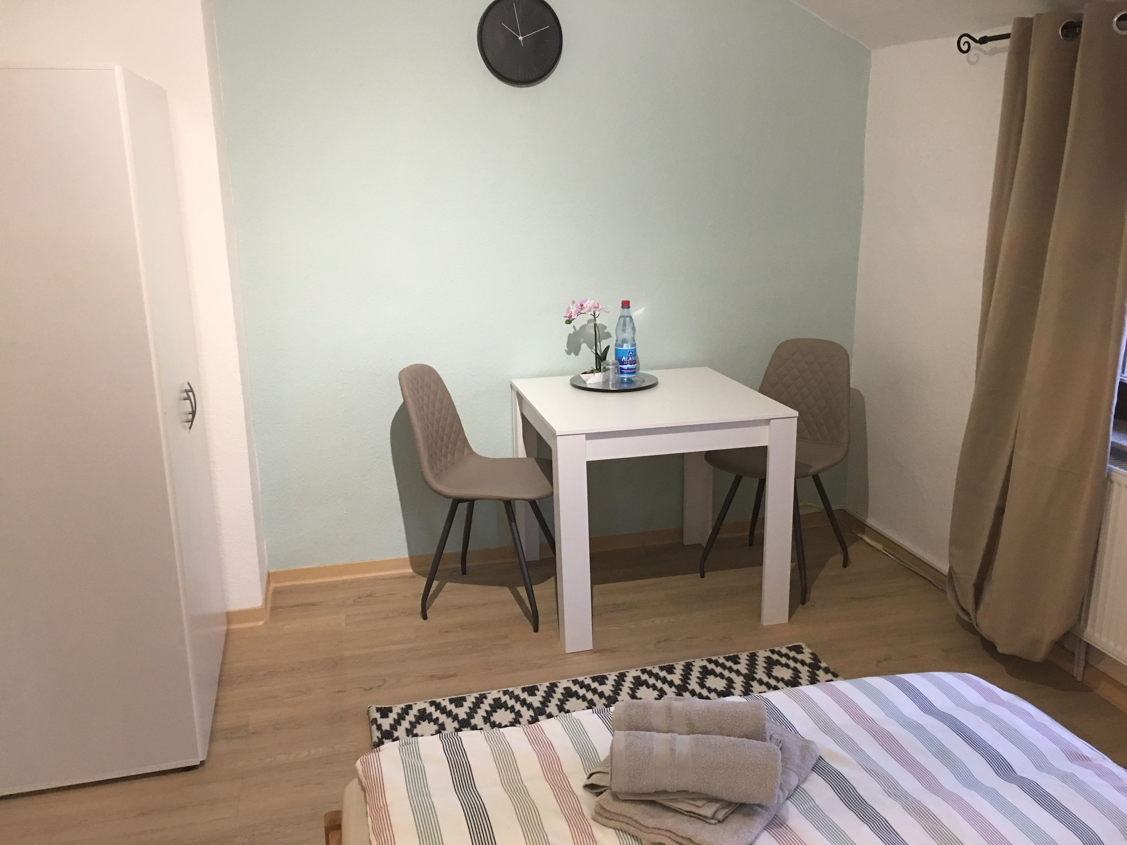 Una habitación pequeña con una cama, una mesa y dos sillas, un reloj en la pared y una ventana con cortinas.