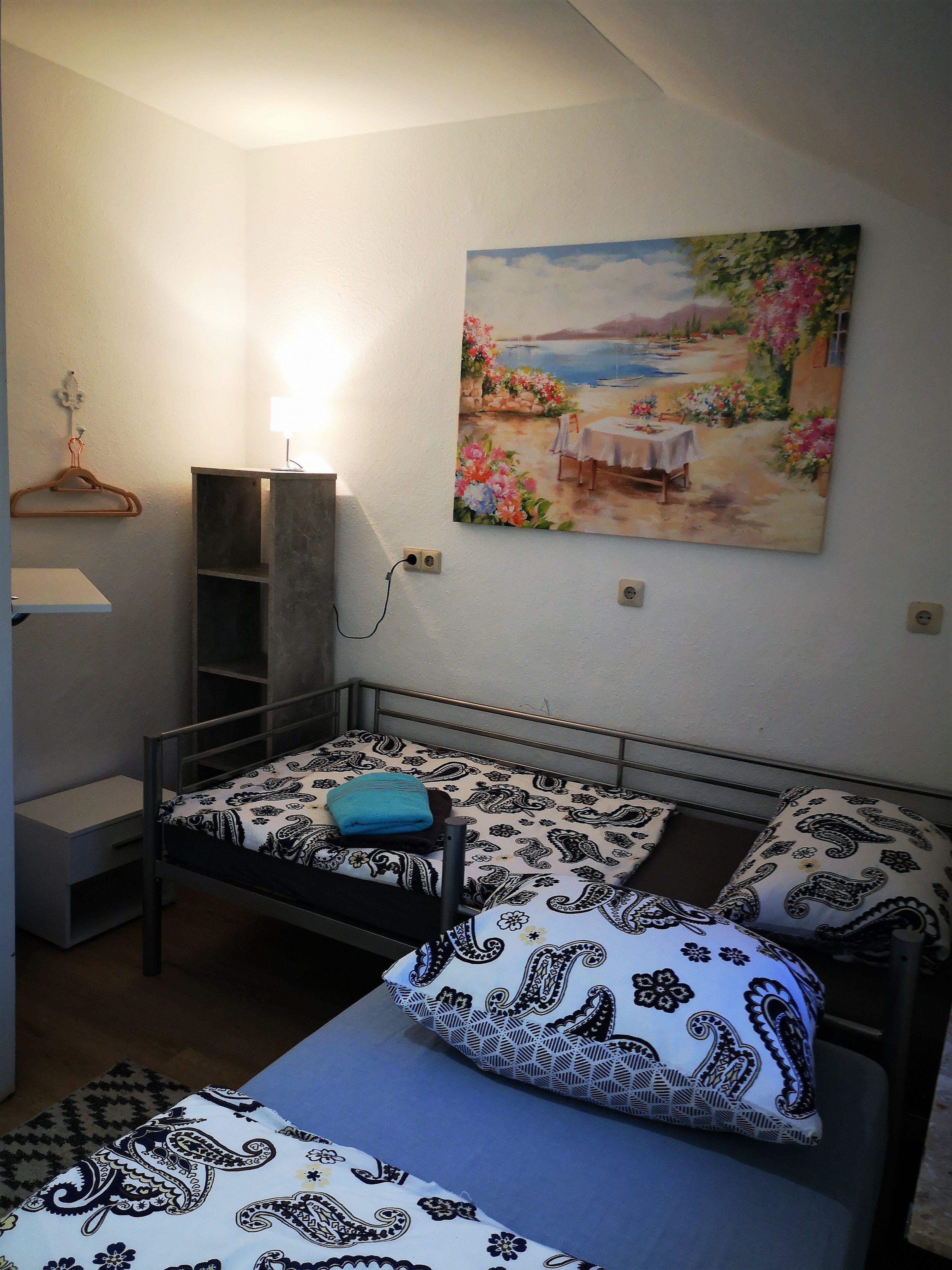 A bedroom with a bed, bedside table, and a painting of a garden scene.