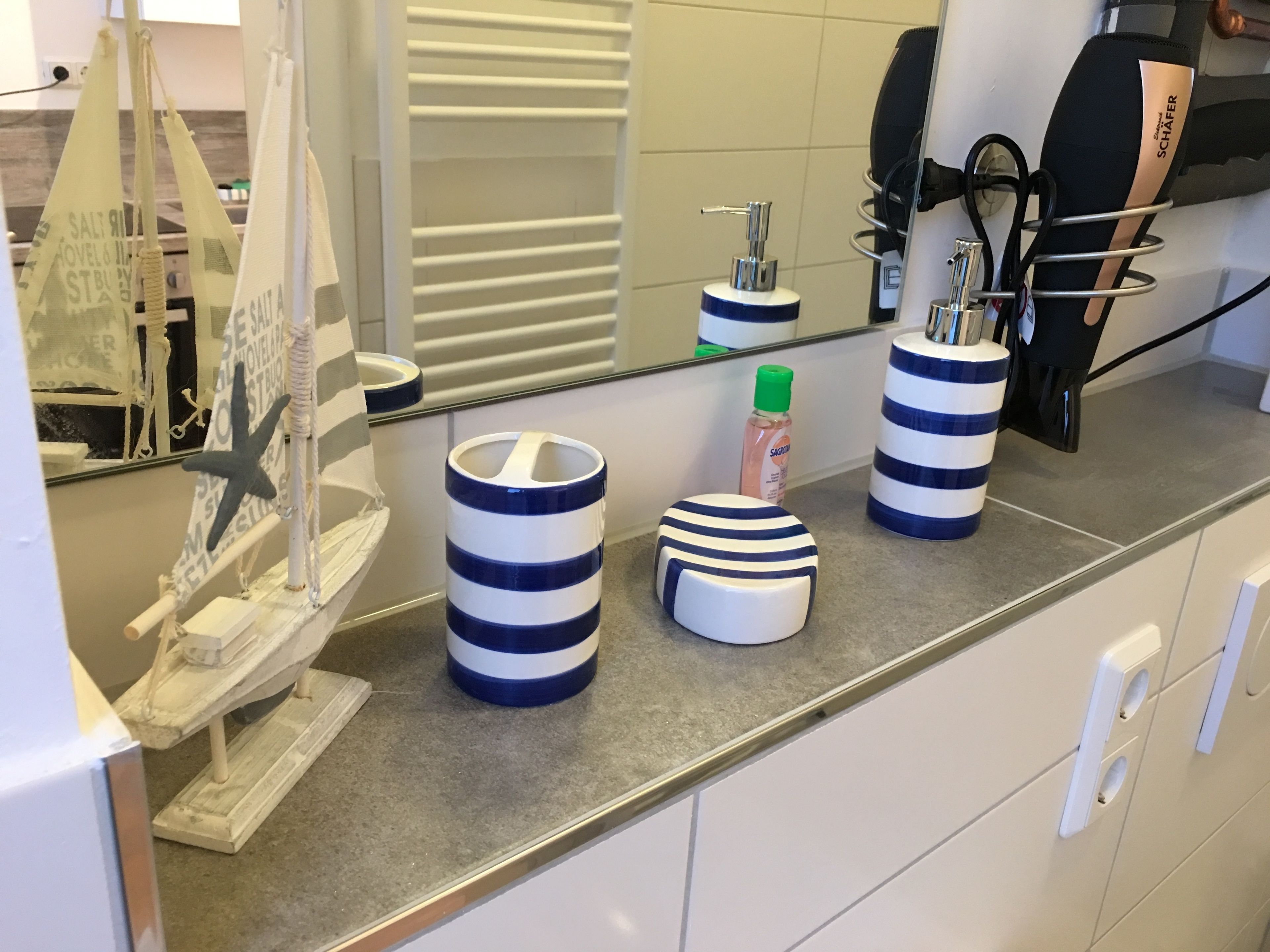 Une salle de bain avec un grand miroir, des distributeurs de savon à rayures, un porte-brosse à dents et une maquette de bateau décorative.