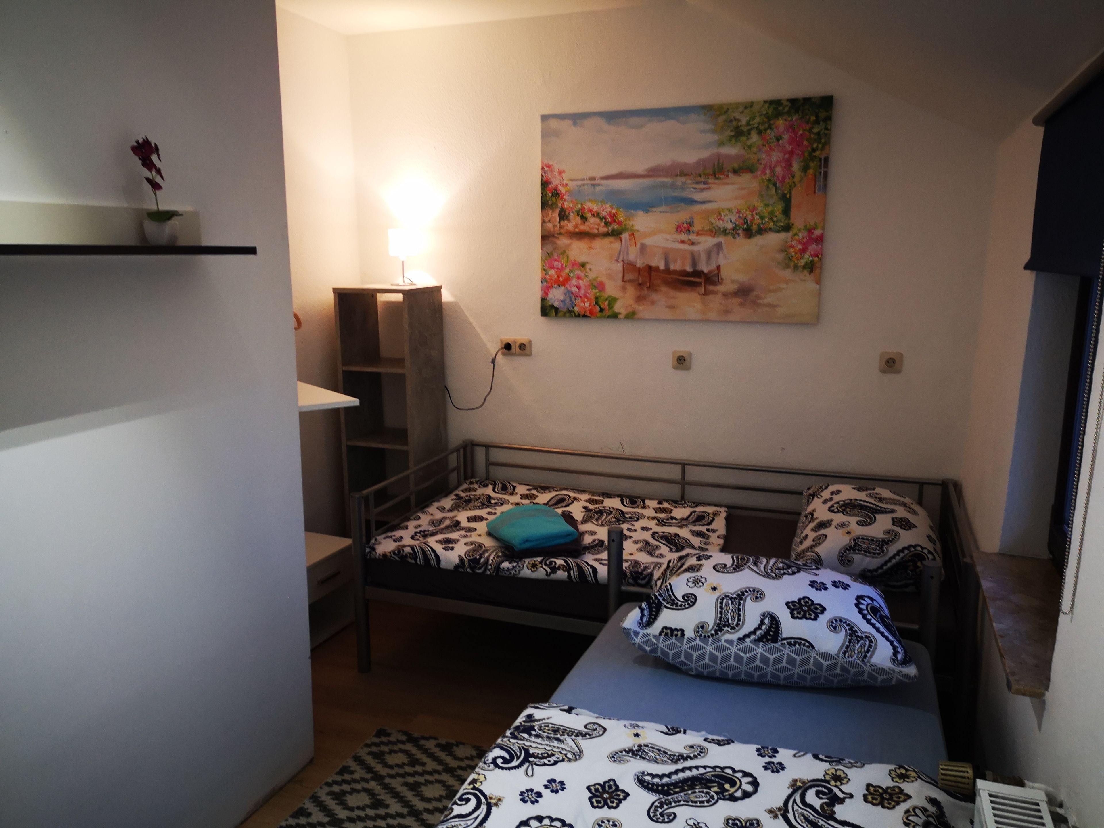 A small, cozy bedroom with a single bed, a nightstand, a lamp, and a painting of a landscape.