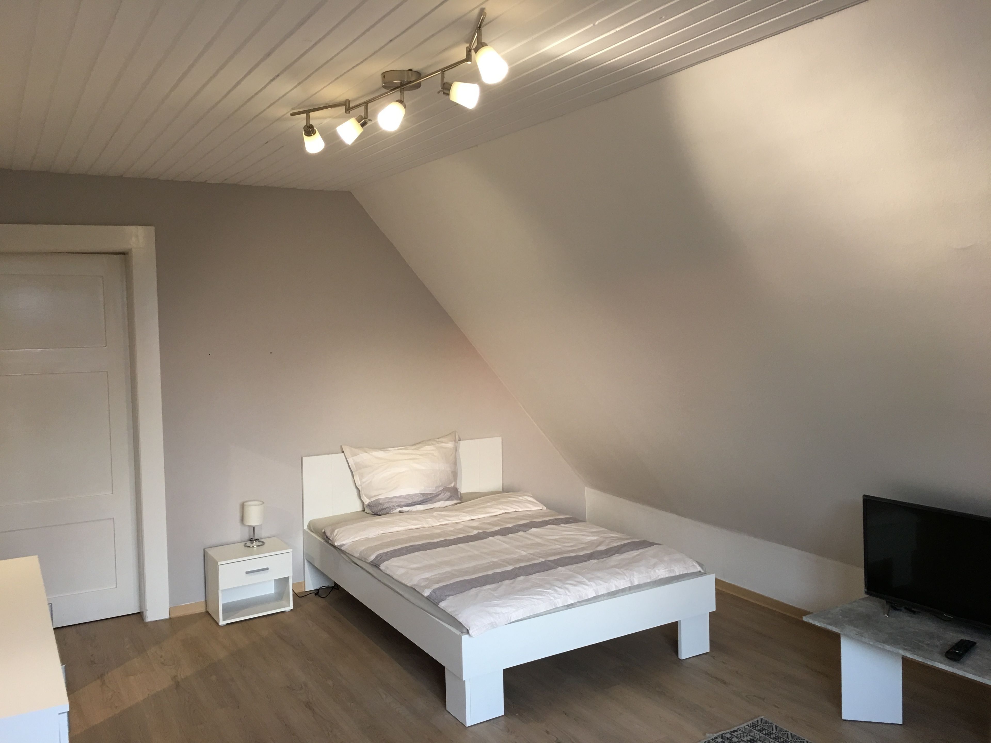 Un dormitorio con techo inclinado, una cama con colcha a rayas, una mesita de noche con lámpara, un televisor sobre un mueble y una puerta.
