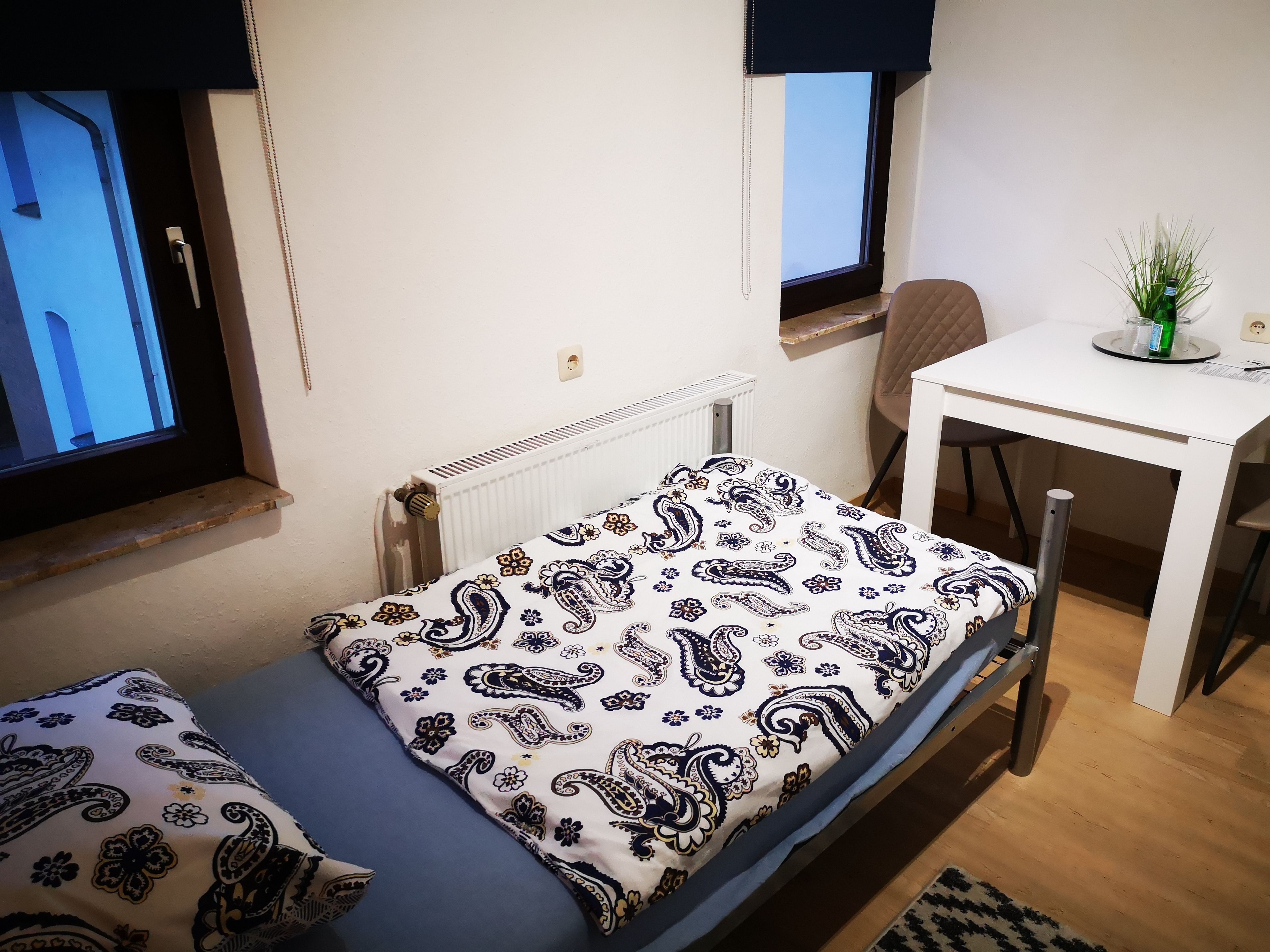 Ein kleines, ordentlich eingerichtetes Schlafzimmer mit einem Einzelbett, einem Sessel, einem kleinen Tisch und einer Heizung.