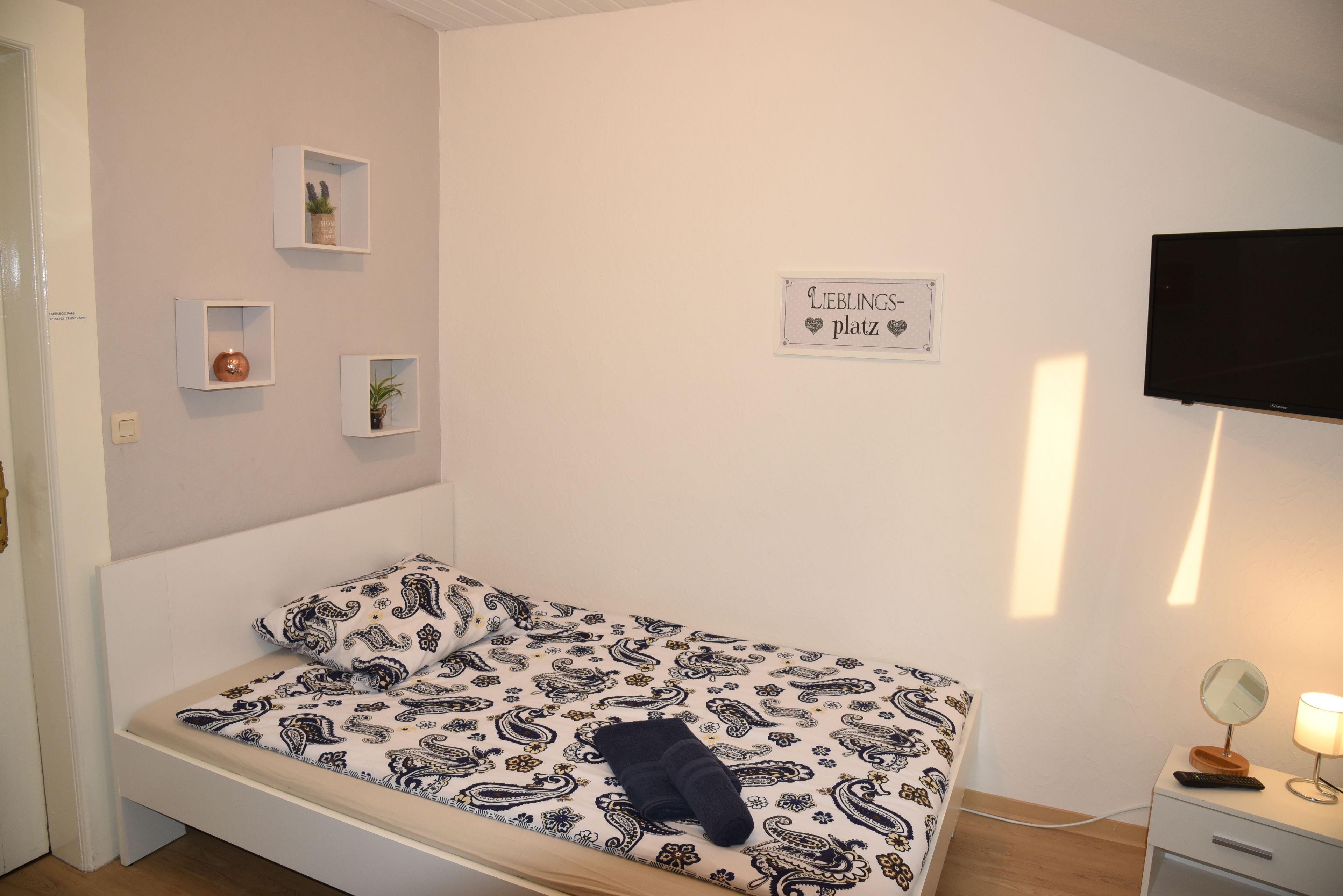 Un dormitorio con cama, una mesita de noche con lámpara, televisión y tres estantes en la pared con objetos decorativos.