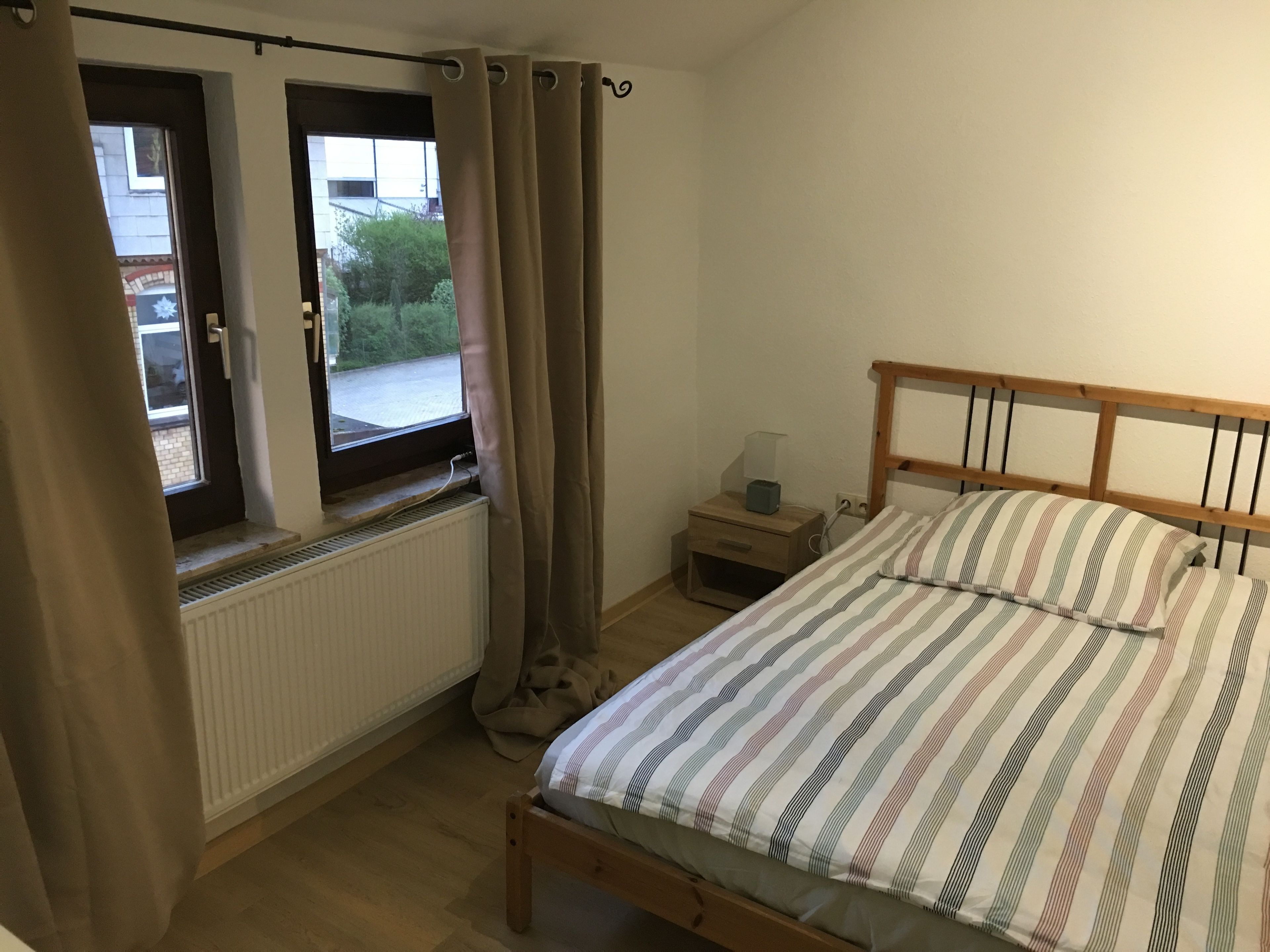 Ein Schlafzimmer mit Bett, Nachttisch, Fenster mit Vorhängen und Heizkörper.