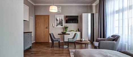A modern hotel room with a wooden floor, a bed, a desk with two chairs, a TV, and a large window with curtains.