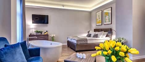 A modern hotel room with a large bed, a sitting area with a sofa and a coffee table, a bathtub, and a flat-screen TV mounted on the wall.