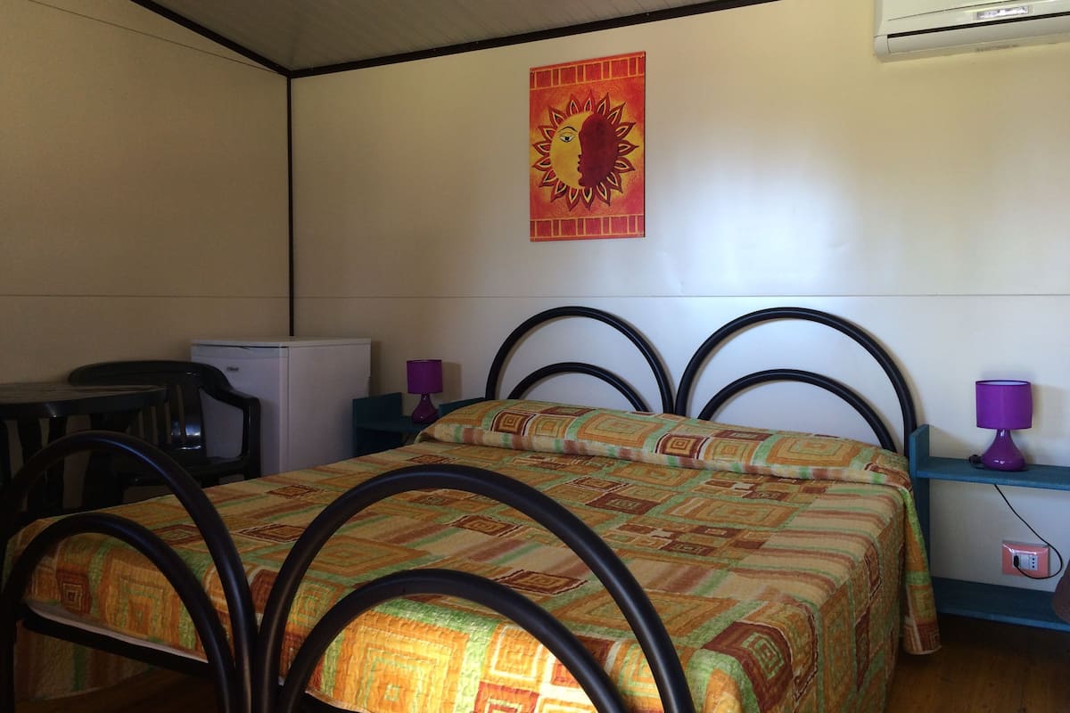 Un lit simple avec une couvre-lit à motifs, un petit bureau avec une chaise, un climatiseur fixé au mur et un élément décoratif mural.