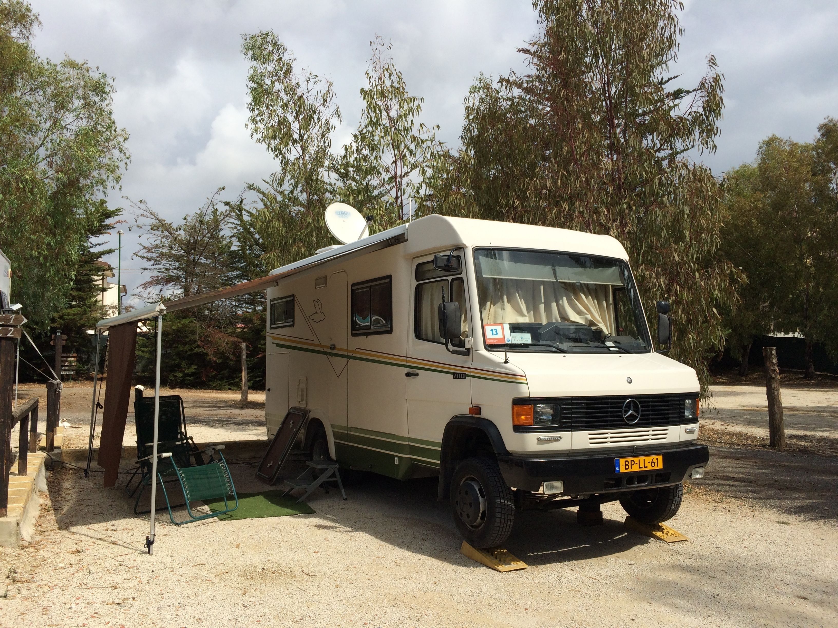 Una camper blanca estacionada en un camping con una antena parabólica en la parte superior, una silla plegable verde y una mesa pequeña.