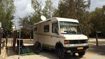 Una camper blanca estacionada en un camping con una antena parabólica en la parte superior, una silla plegable verde y una mesa pequeña.