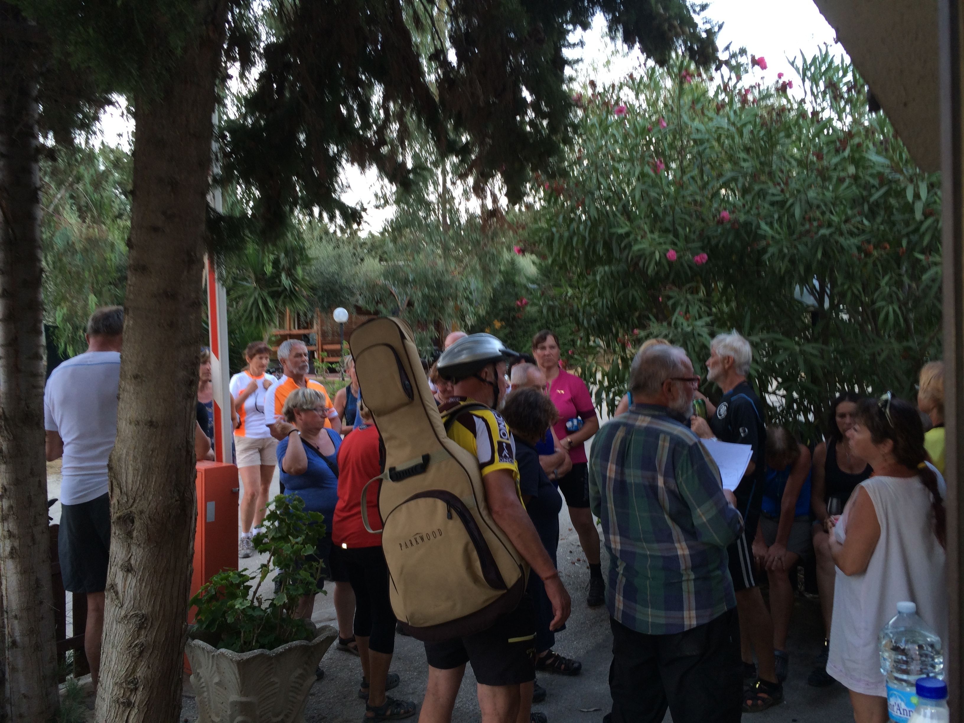 A group of people gathered outdoors, some holding documents, in a shaded area with trees and plants.