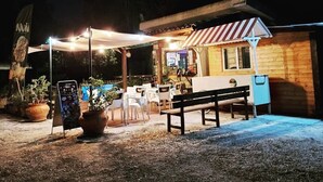 An outdoor seating area with tables and chairs, a wooden building with a striped awning, and a signboard. - Camping Valle Dei Templi (Agrigento)