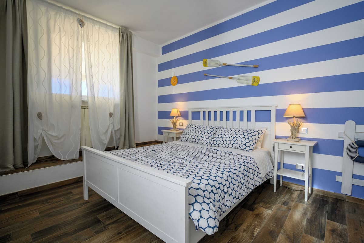 Un dormitorio con una cama, dos mesitas de noche con lámparas, una ventana con cortinas y una pared a rayas azules y blancas.