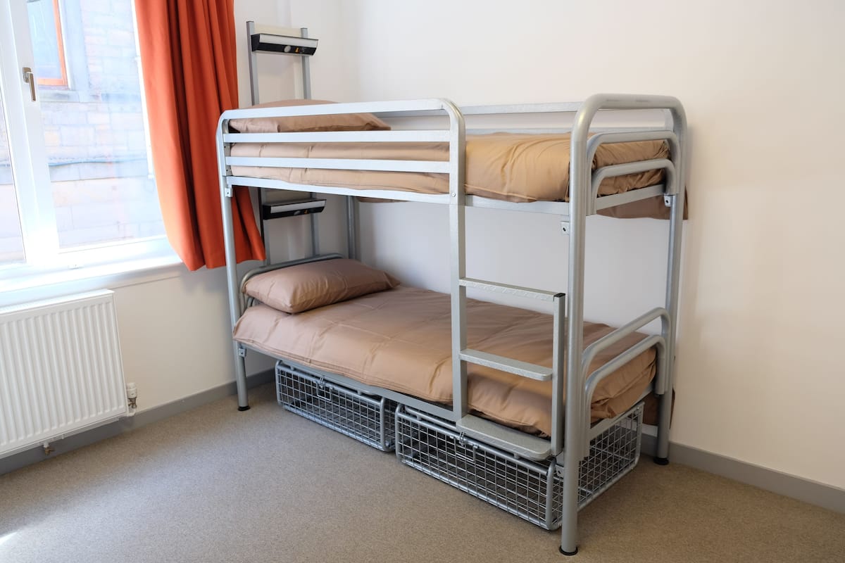 Una cama litera con tres niveles, una ventana con cortinas y un radiador.