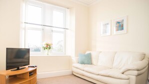 A living room with a beige sofa, a television on a wooden stand, and a window with a view of trees.