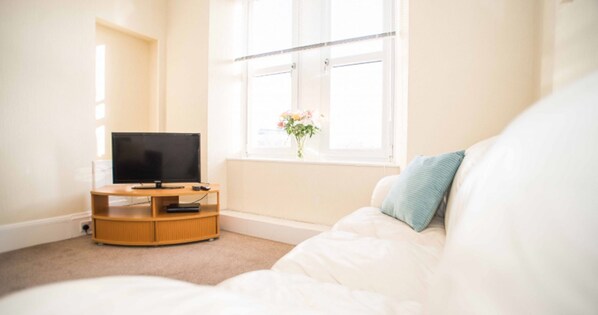 A room with a bed, a TV on a wooden stand, and a window with a flower vase.