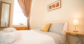 A neatly made bed with a pillow, a bedside table with a lamp, a window with curtains, and a framed picture on the wall.
