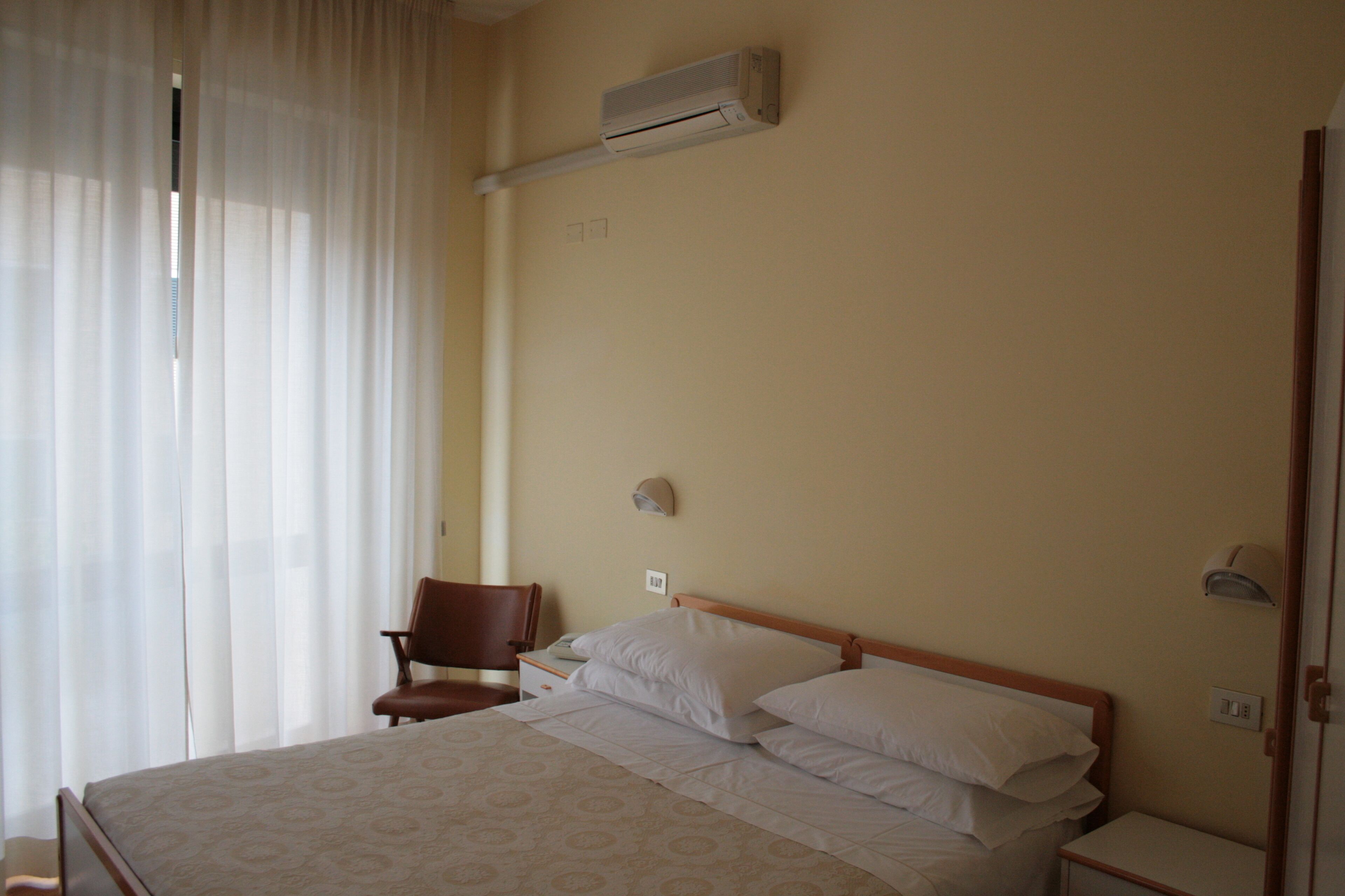 A hotel room with a bed, a chair, a nightstand, and an air conditioning unit.