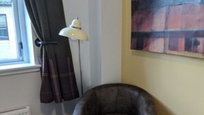 A small, dark leather armchair in a corner of a room with a radiator, a floor lamp, and a window with curtains. - Black Isle Bar and Rooms (Inverness)