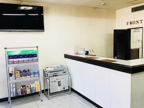 Bancada de recepção com placa 'FRONT', televisão montada acima e um suporte de revistas com diversas publicações.