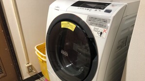 Una lavadora de carga frontal blanca con puerta negra, un cubo amarillo y un timbre cerca de un suelo de baldosas.
