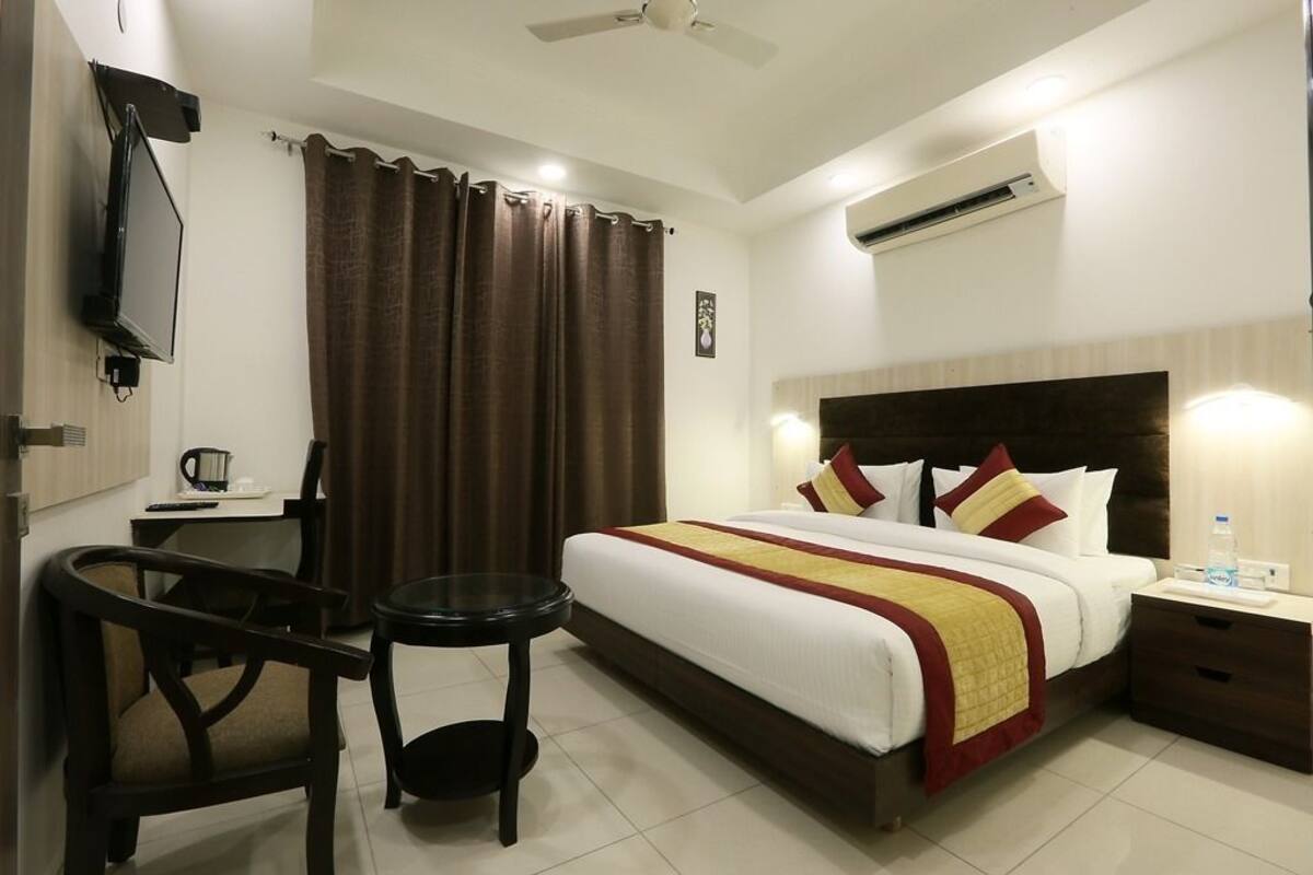 A hotel room with a bed, bedside table, chair, TV, and an air conditioning unit.