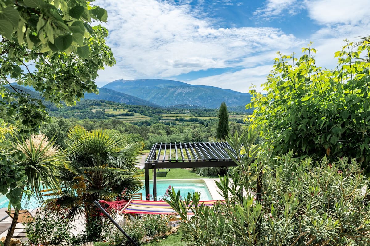Zona de piscina amb una pèrgola, envoltada de vegetació exuberant i un paisatge de muntanyes al fons.