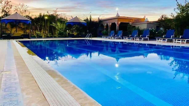 Una piscina cristalina con sillas de descanso y sombrillas, rodeada de vegetación y edificios.