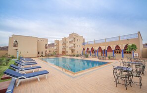 A pool area with lounge chairs and tables, surrounded by a building with arches and a covered walkway. - Ksar Ben Youssef (Tarmigt)