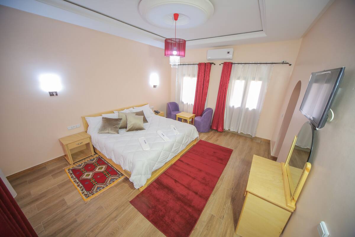 Quarto com cama, mesas de cabeceira, tapete vermelho, cadeira, mesinha e televisão.