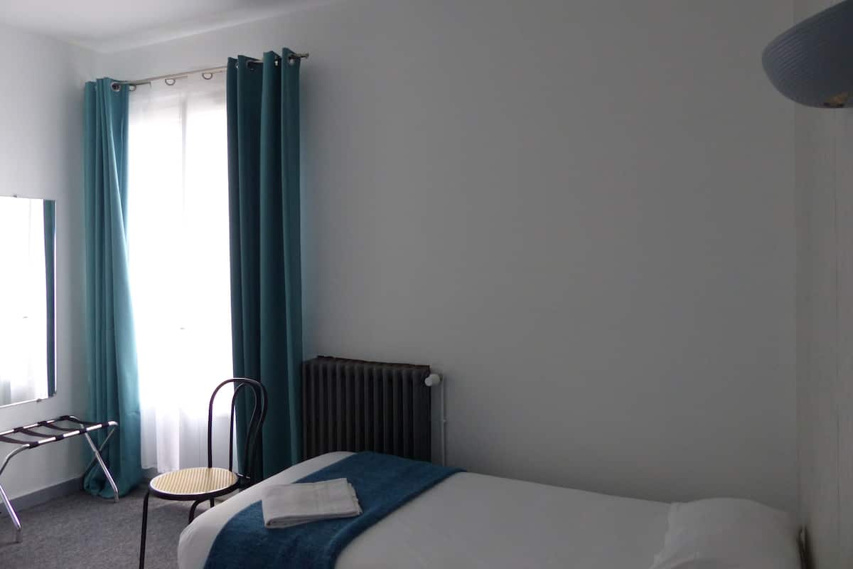 A hotel room with a bed, a chair, a radiator, and a window with curtains.