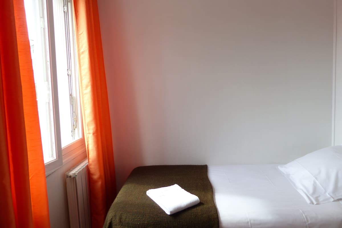 A small, single-bed room with a window, a radiator, and a white towel on the bed.