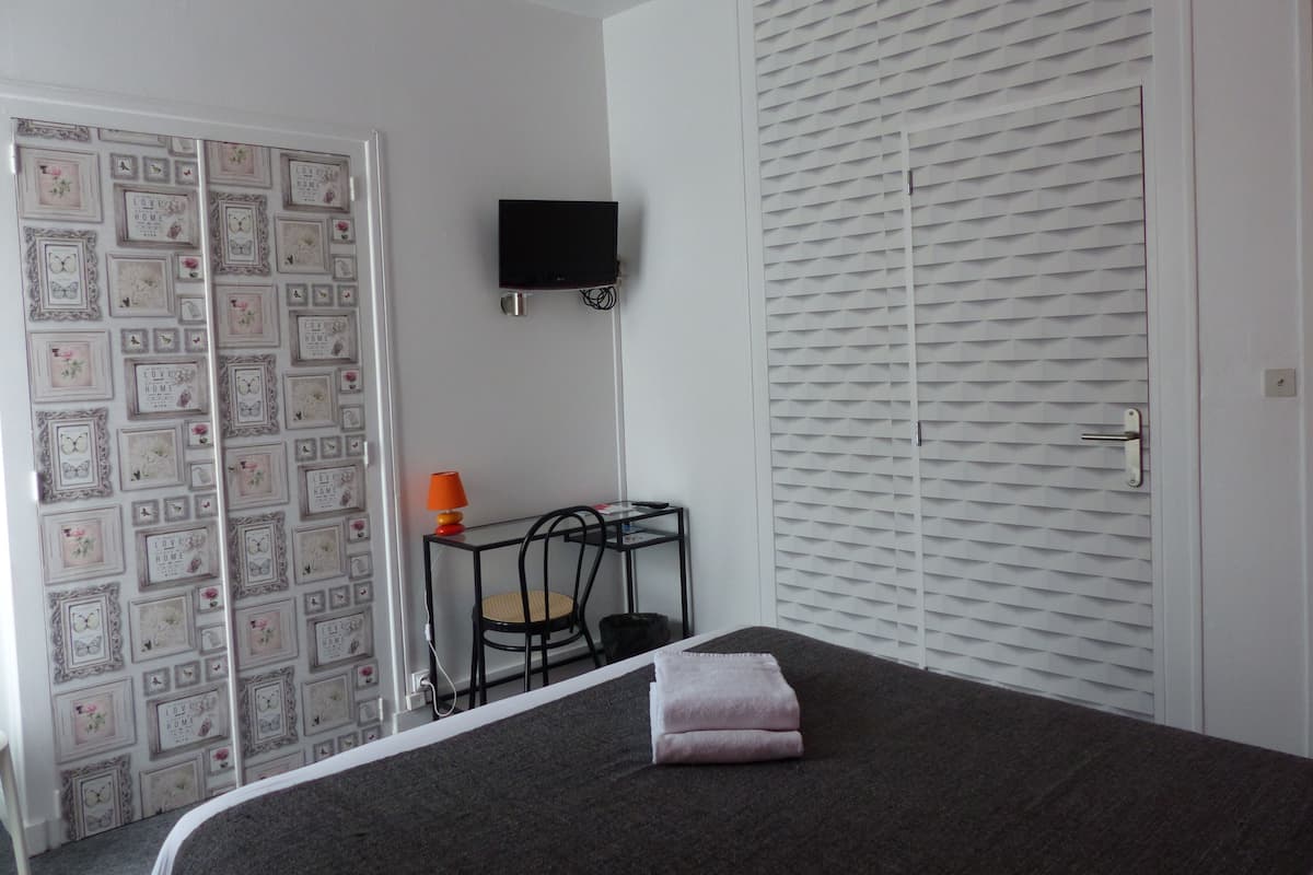 A bedroom with a bed, a TV mounted on the wall, a wardrobe, and a small table with a lamp.