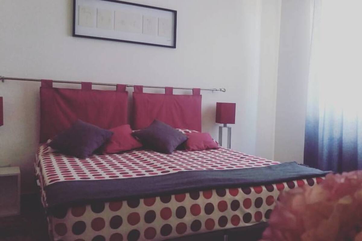 A bedroom with a bed, red curtains, a framed picture on the wall, and a window with a view.
