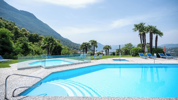 A pool area with clear blue water, surrounded by palm trees and a mountainous backdrop. - Centro Cadro Panoramica (Lugano)
