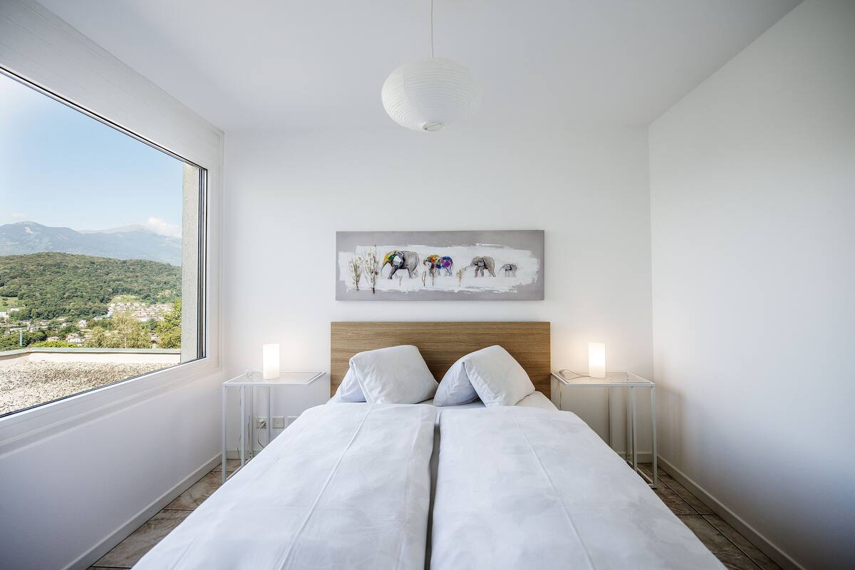 A bedroom with a large window, a wooden headboard, a painting, and bedside lamps.